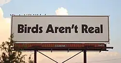 A billboard reading "Birds Aren't Real" in large black font on a white background. The billboard has several small alleged pigeons perched on top of it.