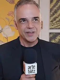Ângelo Antônio Carneiro Lopes wearing a black t-shirt under a black suit jacket, speaking into a microphone and looking left of camera