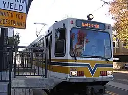 A white and yellow light rail train at a station platform with overhead lines visible above.