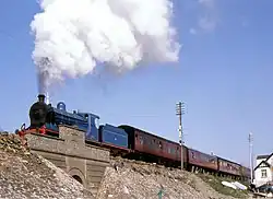 A blue steam locomotive pulling at least four brown passenger coaches over a small bridge; a beautiful plume of white smoke erupts from her funnel.