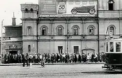 Black and white photograph of a three-floor façade. On the top floor a large poster has Cyrillic text and a picture of a roofless car in profile and several rectangles labelled "50 коп.". On the street, people queues towards a gate. A passing tramway covers part of the queue.