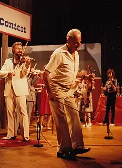 A man in a suit fiddles on stage behind a man holding a fiddle while dancing in front of him.