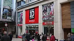A "1D World" shop in England. Above the shop's entrance is a bright red banner with One Direction's logo on it. On the sides of the shop are images of the members of One Direction together. The image is taken on a street, and there are several people walking past the shop.