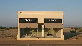 A small one story, one room adobe building with two large plate glass windows. Purses and shoes are sparsely and methodically placed inside.