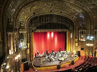 The proscenium and stage of the United Palace. The stage is set up for an orchestral performance, with band equipment such as drums on stage. The proscenium arch is elaborately decorated.
