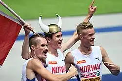 Photo of Henrik, Jakob, and Filip Ingebrigtsen, where Jakob is wearing a viking helmet with two horns and carrying a short flagpole with a Norwegian flag partially visible