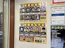 Photograph of two posters on a wall depicting mugshots of wanted Japanese criminals. One of the posters contains Satoshi's mugshot, in which he is grinning.