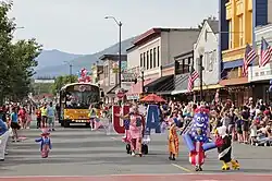 View of a parade with spectators lining a small town street observing children dressed as a bald eagle, Uncle Sam, and other characters, followed by an adult in a "USA" outfit and a school bus.