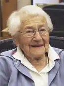 Photograph of Dr. Lucy Ozarin from 2012. She is smiling, while sitting at a desk with a few papers on the desk. She is wearing a white collared shirt and a blue jacket.