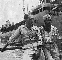 Black and white photograph of two people in khakis standing near a railing with a ship in the background