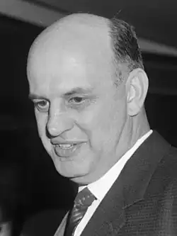 A black and white photo of a white man with thinning hair, wearing a suit and smiling.