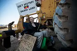 A woman sits with a chain around her waist, padlocked to heavy earthmoving equipment.