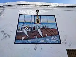 Photo from below of a white wall featuring 8 by 10 painted tiles. On the tiles, a boat or boats crewed by 11 white men. Some of the men are hauling two big tuna onto the boats. Brown nets cover the side of the boats into the water. Three white men on the net are partially into the water gesturing to the men on the boats. 6 tuna swim on the water near the nets. On the center top of the tiles there is a rectangle with a picture of a standing crowned woman holding a child and a scapular.
