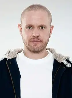 frontal image of Anders Matthesen wearing a white t-shirt and black jacket, staring directly at camera