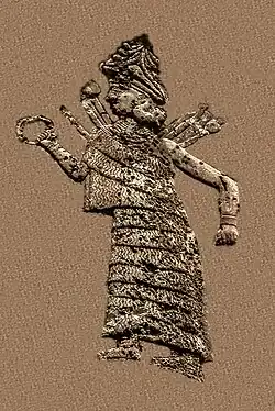 Goddess Ishtar/Inanna. She wears a long, flounced dress, a hat decorated with horns and a headed collar. She is extending a ring in her right hand and has club-like weapons in her back.[1]