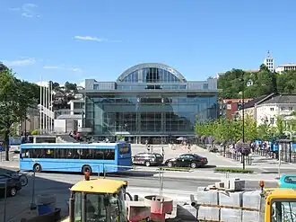 The front of Arendal Town Hall, primarily made of glass. A road in front of the Hall has parked cars and a bus.