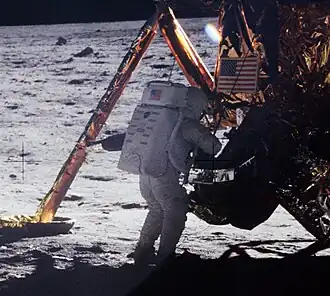 A grainy picture from behind of a human figure in white space suit and backpack standing in front of the Lunar Module on the surface of the Moon. A landing leg is visible and the U.S. flag on the descent stage.