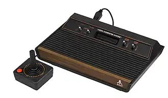 Photograph of a black flat rectangular plastic video game console with brown trims. The top rear portion of the device is raised with a receptacle slot for game cartridges flanked by two sets of two silver switches. A black cable connects to the back of the console and wraps around the left side to black flat square plastic game controller with a red button and a moveable stick protruding upwards.