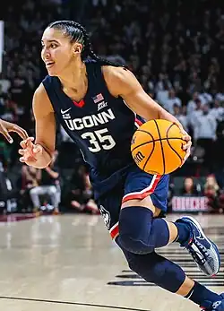 A basketball player, wearing a navy blue uniform, dribbling the ball with her left hand.