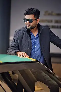 William standing, wearing sunglasses and a vest with his hand resting on the roof of a car in front of him