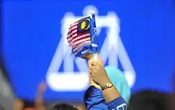 A raised hand holding a Malaysian flag and a Barisan Nasional flag in front of a large Barisan Nasional flag