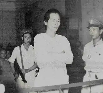 A dishevelled man dressed in white folds his arms as he stands in the dock. He is guarded by two soldiers, one with a submachine gun. The courtroom is crowded.