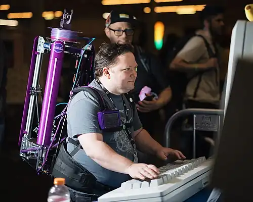 A man wears a back-mounted 3D printer while operating a 237% scale model of a 1986 Macintosh Plus at Open Sauce