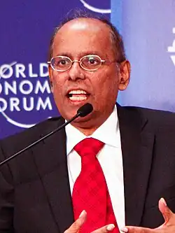 A middle-aged Indian man wearing a black suit, white shirt and red tie speaking into a microphone.