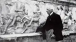 Black-and-white photograph of an elderly man in a dark suit, in front of a wall of Ancient Greek sculptures.