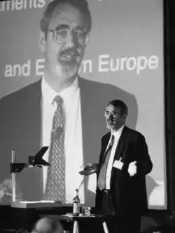 Bearded caucasian male, in his fifties, wearing a suit making a speech; his image is projected on a large screen behind him