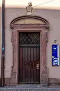 An old wooden door surrounded by a sandstone wall. A blue university of freiburg plaque is on the wall next to it, indicating it is a university building. The door appears accessible, though heavy to open.