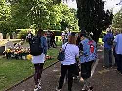 People wearing clothes with Brian Jones and Rolling Stones fan art stand by a gravestone decorated with many flowers in front. One person is placing a bouquet.
