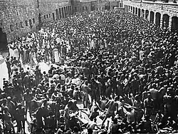 A group several hundred naked men is crowded in an enclosed courtyard, with garage doors visible on three sides.