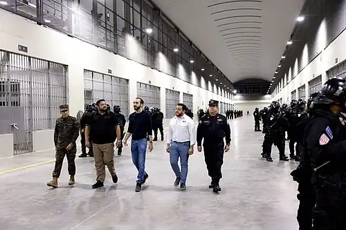 Minister of Defense René Merino Monroy, Director of Penal Centers Osiris Luna Meza, President Nayib Bukele, Minister of Public Works Romeo Rodríguez, and Director of the National Civil Police Mauricio Arriaza Chicas touring a cell block at CECOT while walking past prison guards wearing riot gear