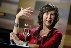 Maria Rita Kehl sitting at a table, in an event, talking ang gesturing with her hands. On the table is a glass of water