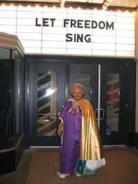 Herron is wearing a purple dress and gold cape, and standing outside a movie theater