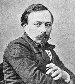 A man with dark receding hair, heavy moustache and a severe expression looks directly at the camera