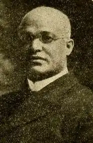 A black man with small, round glasses in a black suit and a high white collar