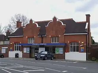 A brown-bricked building with a black roof and a blue sign that reads "CHIGWELL STATION" in white letters all under a white sky