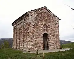 (Top image): A worn red brick building with decorative inlets on the side; the roof of the building is a gable roof; the door is a highly weathered wooden door with a decorative indent just above that is in the shape of a window. (Church of Saint John Orljane) (Bottom Image): Map of Serbia with the location of the settlement, in south eastern Serbia, marked by a red dot.