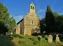 Medium size stone building with pitched roof, arched entrance door and a short tower having exposed double bells on a sunny day against partly overgrown graveyard to front and blue sky beyond