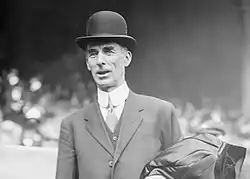 A man standing in a baseball stadium wearing a suit and bowler hat. He has a coat folded over his left arm.