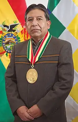 Full body portrait of David Choquehuanca wearing the Vice Presidential medal, flanked by the Bolivian flag on the left and the Wiphala on the right.