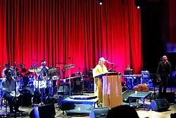 An ensemble are performing on-stage, three musicians are seated at extreme left behind musical instruments. Lisa Gerrard is behind a lectern near mid-stage with a microphone. At the right is Brendan Perry holding a microphone with his left hand. His right hand is alongside his thigh and holding an instrument. The background includes a long stage curtain with another musician seated at rear right, who is obscured behind a keyboard.