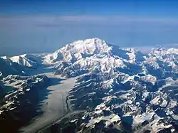 In an aerial image, a mountain is surrounded by many smaller mountains and a glacier