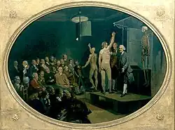 A man stands on a stage with a skeleton, an anatomical model, and a man stripped to the waist. A crowd of seated men are watching.