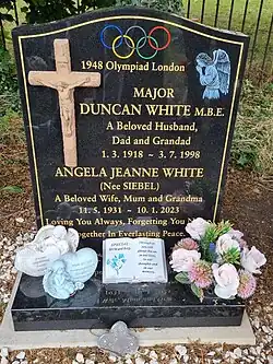 A memorial headstone headed with the Olympic wings, featuring a crucifix and an angel, and reading "MAJOR DUNCAN WHITE M.B.E. A Beloved Husband, Dad and Grandad 1.3.1918 to 3.7.1998. ANGELA JEANNE WHITE (Nee SIEBEL) A Beloved Wife, Mum and Grandma 11.5.1931 to 10.1.2023. Loving You Always, Forgetting You Never. Together In Everlasting Peace."