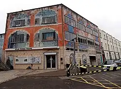 A wide three storied building with windows to the upper two stories and an entrance on the ground floor. A statue of Thomas sits outside.