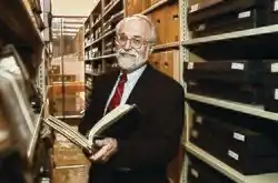 A photograph taken in November 2010, of then Maryland State Archivist and Commissioner of Land Patents, Dr. Edward C. Papenfuse, holding a book, possibly which are bounded records, while standing with a smile on his face, in a row of shelved stacks of other bounded books, in the archive.