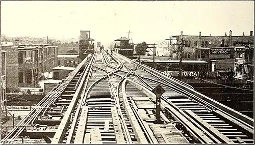 A sepia image of a double-tracked elevated line with a diamond crossing near the camera. In the distance is a station with two platforms with hipped-roof canopies, with the left platform having a square tower.